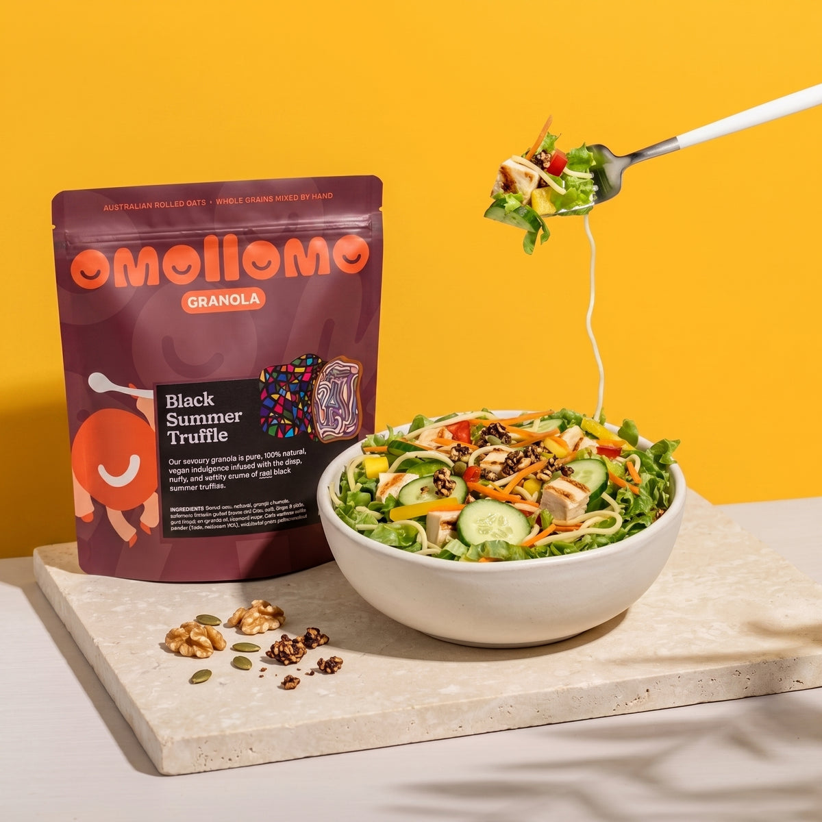 Omollomo Black Summer Truffle Savoury Granola pouch standing on a stone slab next to a colorful salad bowl. A fork lifts a bite of salad against a vibrant yellow background with soft leaf shadows in a clean, commercial style.