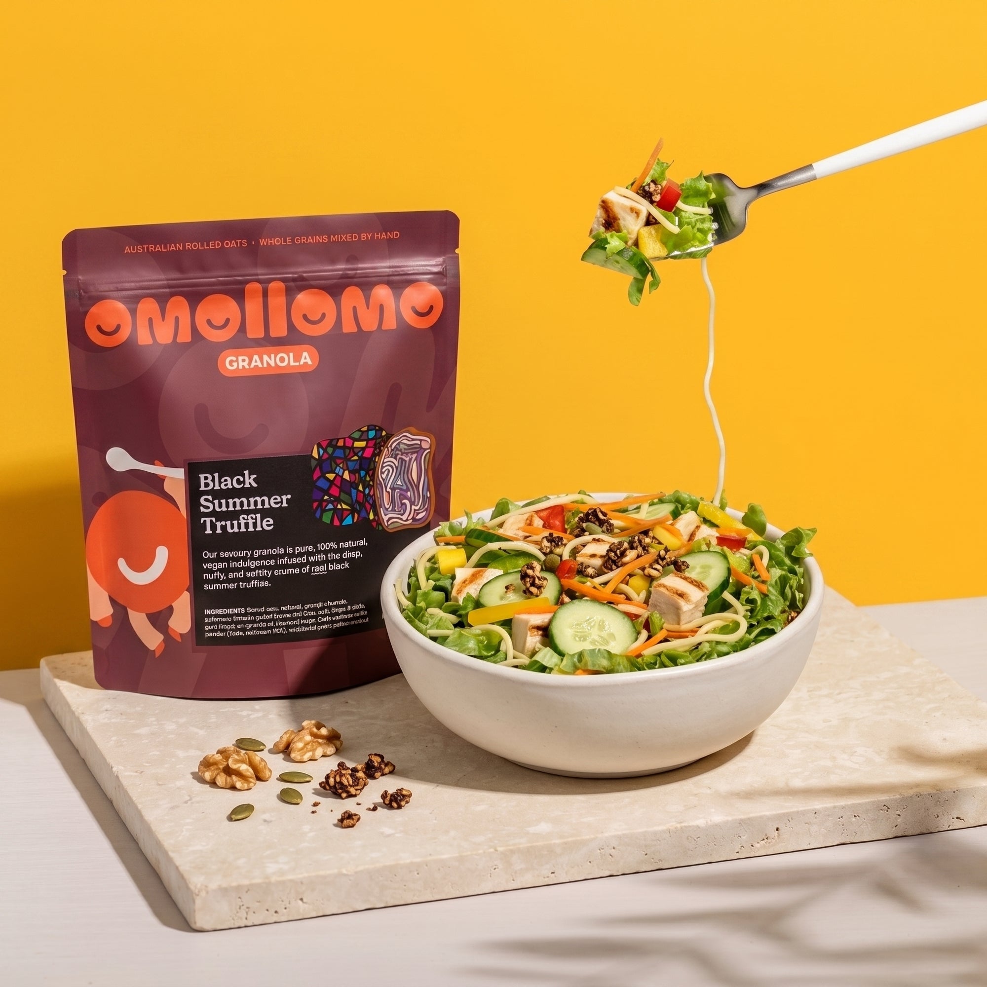 Omollomo Black Summer Truffle Savoury Granola pouch standing on a stone slab next to a colorful salad bowl. A fork lifts a bite of salad against a vibrant yellow background with soft leaf shadows in a clean, commercial style.