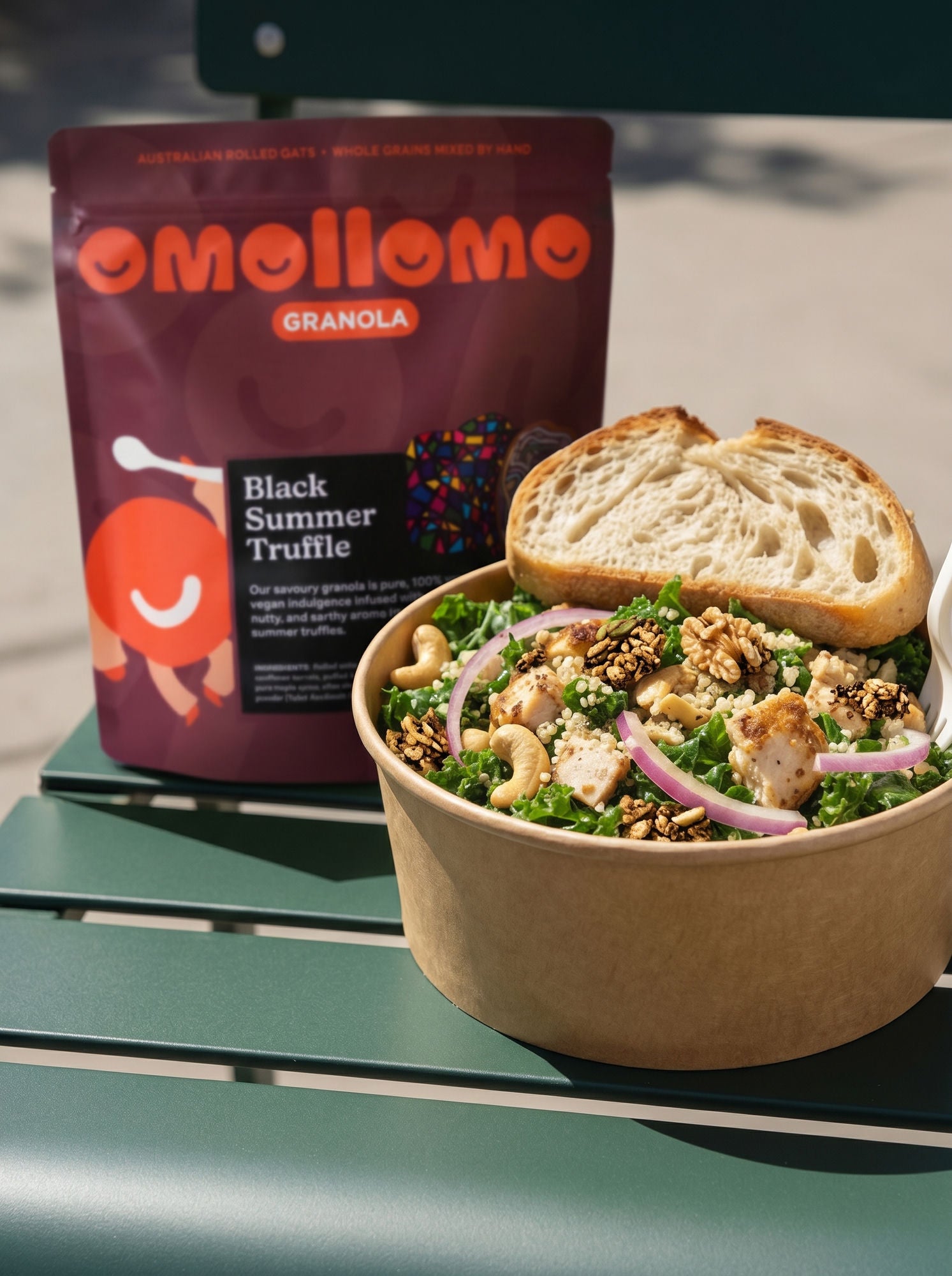 Omollomo Black Summer Truffle Savoury Granola pouch beside a kale, grain and chicken salad bowl with sourdough